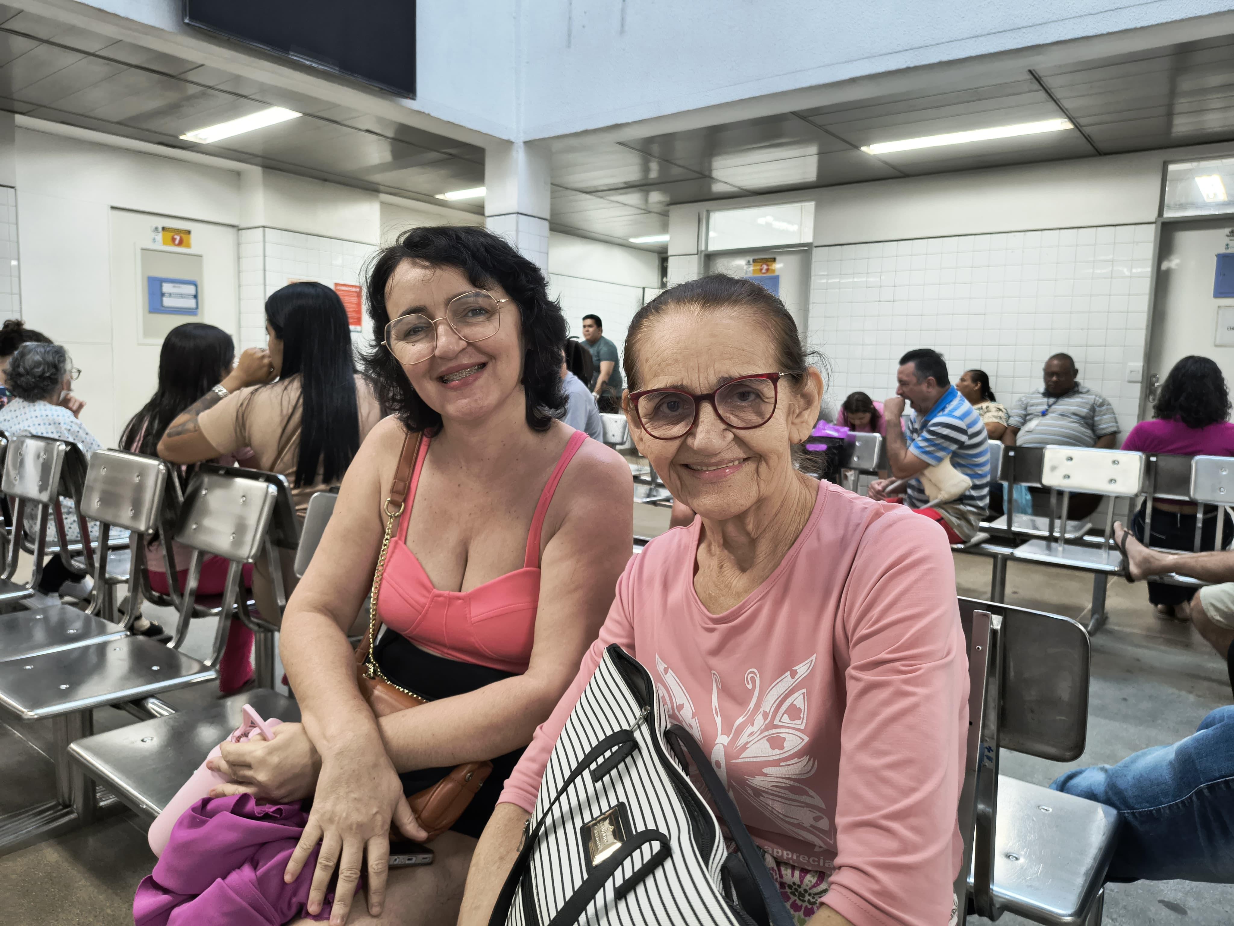 duas mulheres posam para a foto, sentadas em cadeiras numa recepção de hospital